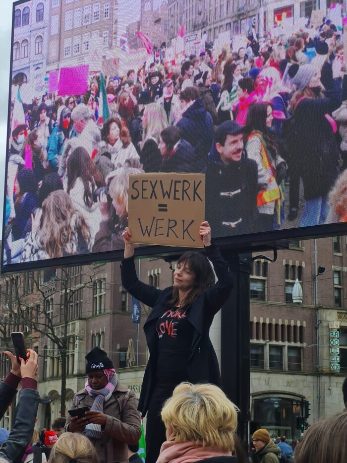 Vrouw houdt tijdens demonstratie op feminist march 2023 bord omhoog: sekswerk is werk. Ze is in het zwart gekleed. Op haar shirt de woorden: Whore Love